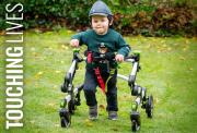 A picutre of Barnaby, a young boy in a green bear sweater and cap smiles while using a supportive walking frame in a grassy garden with fallen leaves