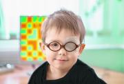 A boy, aged about two or three, wearing brown-rimmed glasses and with an eye patch over his right eye. The background is a pale green wall with a blurred window.  