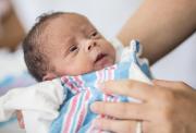 A newborn baby, with black hair, in the hands of a caregiver in a hospital setting. The baby is wrapped in a blue, pink and white stripe sheet or blanket.