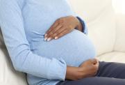 Picture of the side view of a pregnant woman, from the shoulders down, sitting on a cream sofa. Her hands are cradling her baby bump and she is wearing a blue top.