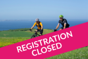 Two mountain bikers riding across a grassy hill overlooking a blue sea, with a large pink diagonal banner in the foreground that reads "REGISTRATION CLOSED" in white capital letters.