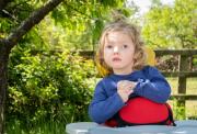 Image shows Emmy, a little girl who has Vici syndrome. She has light brown wavy hair and wears a blue top. She is using supportive seating with a play tray in front of her. She is outside with trees, bushes and a wooden fence in the background. 