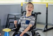 Image shows a smiling boy, aged around 12 or 13. He wearing a striped t-shirt and is in his wheelchair, with exercise equipment in the back and foreground
