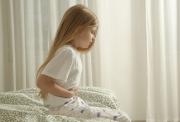 A young girl holding her stomach in pain. She has long, blonde hair and is sitting on the end of a bed, wearing pyjamas. There are cream-coloured curtains in the background.