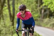 A close-up panoramic view of a female cyclist wearing a helmet, sunglasses, and a pink and blue Fdx jersey with trees behind her.