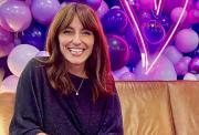 Image shows Davina McCall sitting smiling on a light brown leather sofa. There is a wall of balloons in purples and lilacs behind her.