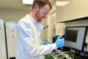 A smiling male, Dr Michael Wilkinson, researcher in a white lab coat and blue gloves uses a pipette next to a computer monitor in a laboratory.
