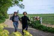 Two hikers smiling as they walk along a dirt path next to a green field and a wooden gate on a clear day.
