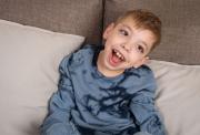 Smiling young boy in a blue tie-dye shirt sits on a couch with beige cushions, looking cheerful and engaged.
