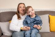 A woman and a young boy sit smiling on a gray couch with yellow and gray cushions. The woman wears a white sweater and jeans, while the boy, who appears to have a disability, wears a blue tie-dye sweatshirt and jeans. They are holding hands and looking happy.