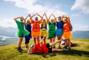 A group of women in colorful, crayon-themed shirts and tutus pose joyfully on a grassy hill overlooking a scenic mountain and lake landscape.