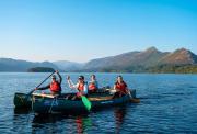 Four people wearing life jackets smiling and paddling in two green canoes side-by-side on a calm lake, with scenic mountains rising in the background under a clear sky.