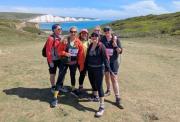 A group of six smiling female hikers posing together on a grassy coastal trail, with a vibrant blue sea and the iconic white chalk cliffs of the Seven Sisters in the background on a sunny day.