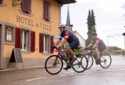 Two men cycling along a damp street in France, beside a beautiful building with the words Hotel de Ville (Town Hall) written on the side
