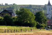 An idyllic German village is the backdrop - in the foreground, a row of cyclists are crossing the edge of a field and waving at the camera