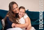 A smiling mother lovingly hugs her young daughter as they sit together on a deep blue velvet sofa.
