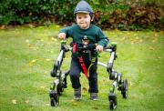 A picutre of Barnaby, a young boy in a green bear sweater and cap smiles while using a supportive walking frame in a grassy garden with fallen leaves