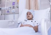 Child in a hospital bed with a bandage around the head, suggesting brain surgery. They are wearing a white gown with black polka dots and resting with their eyes closed.