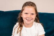 Hattie, a smiling young girl with light brown hair wearing a white top sits in front of a blue velvet couch.