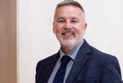 Headshot of a white man with light grey beard and short hair wearing a suit