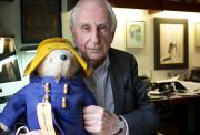 Michael Bond sitting in his study holding a Paddington Bear toy dressed in a yellow hat, blue coat, and red boots.