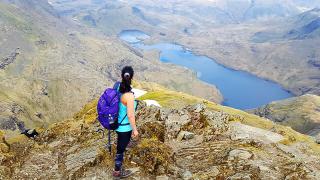 A solo hiker at the summit of Snowdon looking down over rugged landscape with a lake in the distance