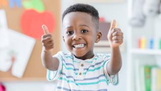 A young toddler age boy smiling at the camera while giving two thumbs up