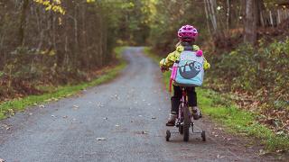 A young child riding their bike with stabalisers on road through the woods alone