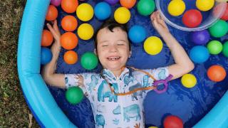 Mylo in a garden lying in a shallow paddling pool surrounded by lots of plastic balls with a big smile on his face