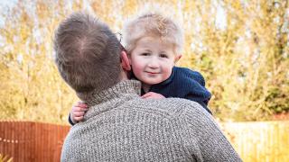 Finely and his father with glasses and a gray sweater hugs his young son with blond, tousled hair, who is smiling and wearing a navy blue sweater.