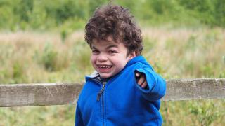  young boy with curly hair wearing a blue jacket, smiling and pointing while standing in front of a wooden fence with greenery in the background.