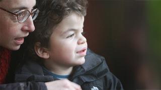 Danny, a young boy with curly hair, wearing a jacket, looks out with a smile while being embraced by his mum in a natural, green setting