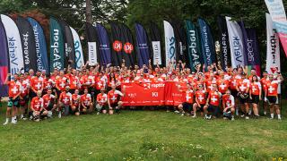 A large group of cyclists in red jerseys with white crosses pose together, holding a red banner, surrounded by sponsor flags in a park.