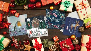 A festive wooden table with Christmas cards featuring winter scenes and decorations on the left, surrounded by gift boxes, pine cones, and ornaments.