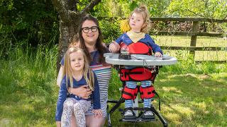 Smiling Mum and sister with along side Emmy standing in a mobility aid, while the other sits on the grass beside her.