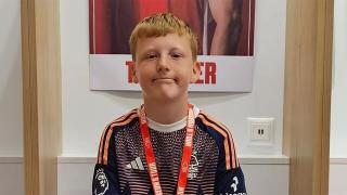 Oscar, a young boy with red hair wearing a sports jersey and lanyard, smiling while seated in a football stadium changing room