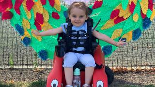 Smiling young girl in a wheelchair, wearing a harness, with vibrant, colorful wing-shaped decorations behind her, arms outstretched