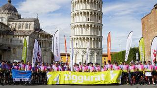 A large group of cyclists in colorful jerseys posing with their bikes in front of the Leaning Tower of Pisa, holding a yellow "Ziggurat" banner