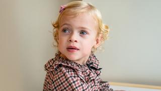 Elsie , a young child with blonde curly hair, wearing a pink bow and checkered outfit, looks to the side with a curious expression