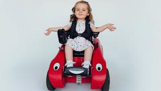 Sophia sitting in a red, child-sized mobility device with cartoon-like eyes, smiling and spreading her arms, wearing a polka dot dress.