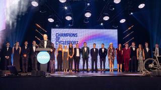 All the cycling champions and ambassadors elegantly dressed standing on stage at an event, "Champions of CycleSport," with dramatic lighting and decor.