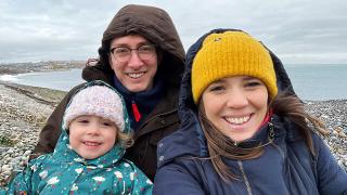 A smiling family of three poses for a selfie on a rocky beach. They are dressed in warm coats and hats, with the ocean behind them.