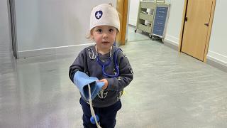  A young child wearing a toy nurse's hat and stethoscope stands in a hospital hallway, holding a play blood pressure cuff.