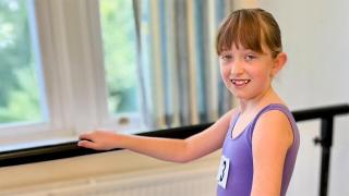  A young girl in a purple leotard stands at a ballet barre, smiling. She wears a number tag, with a bright window behind her.