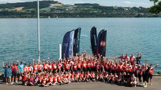 A large group of cyclists in matching red and white jerseys pose with raised hands near a waterfront, with event banners in the background.