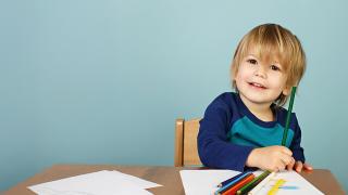 Smiling young child sits at a table holding a green colored pencil, surrounded by blank paper and crayons, ready to draw.