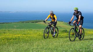 Two cyclists ride mountain bikes along a grassy trail overlooking a scenic coastal landscape. Bright yellow wildflowers dot the foreground, with rolling green fields and a deep blue sea stretching to the horizon under a clear, sunny sky.