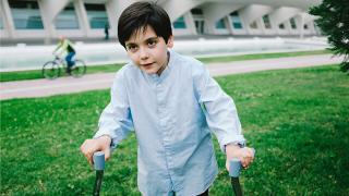 A young boy with dark hair uses forearm crutches to walk on a grassy area. He wears a light blue shirt and looks determined. In the background, a person cycles past a modern building with arched structures and a long reflecting pool.
