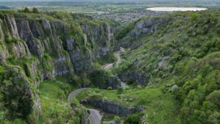 A winding road cuts through the lush, green cliffs of Cheddar Gorge, surrounded by steep rocky walls and dense vegetation.