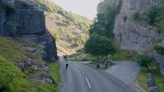 Cyclists ride through a winding road surrounded by steep rocky cliffs and lush greenery in a scenic gorge during daylight